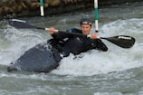 A person wearing a helmet and protective gear is engaged in kayaking, maneuvering through rough, white-capped waters. The paddle is firmly gripped, and the kayak tilts slightly as it navigates through the current, with water splashing around.
