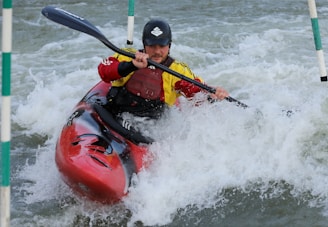 A kayaker navigates through choppy whitewater while holding a black paddle. The person is wearing protective gear, including a helmet and a life jacket, as they skillfully maneuver the red kayak through fast-moving water.