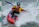 A kayaker navigates through choppy whitewater while holding a black paddle. The person is wearing protective gear, including a helmet and a life jacket, as they skillfully maneuver the red kayak through fast-moving water.