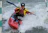 A kayaker navigates through choppy whitewater while holding a black paddle. The person is wearing protective gear, including a helmet and a life jacket, as they skillfully maneuver the red kayak through fast-moving water.