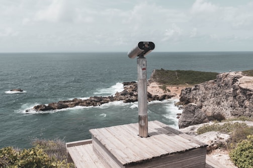 A scenic coastal landscape featuring a telescope mounted on a wooden platform overlooking a rocky shoreline. The ocean extends into the horizon, with gentle waves hitting the rugged rocks below. Sparse green vegetation covers the cliffs, and the sky is partially cloudy.