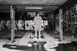 A person stands in an industrial setting surrounded by graffiti-covered walls. The space appears to be an abandoned or disused garage or workshop area, with a concrete floor and an overhead structure. The lighting is dim, casting shadows throughout the scene, creating a gritty, urban atmosphere.