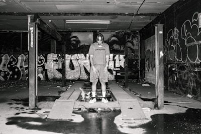 A person stands in an industrial setting surrounded by graffiti-covered walls. The space appears to be an abandoned or disused garage or workshop area, with a concrete floor and an overhead structure. The lighting is dim, casting shadows throughout the scene, creating a gritty, urban atmosphere.