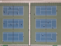 An aerial view of six tennis courts arranged side by side with players actively engaged in games. The courts are separated by fences and netting, and shadows indicate the presence of bright sunlight.