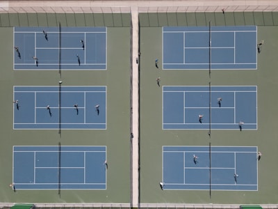 An aerial view of six tennis courts arranged side by side with players actively engaged in games. The courts are separated by fences and netting, and shadows indicate the presence of bright sunlight.