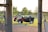 A utility vehicle with a red and black body is parked on a grassy field, near a baseball diamond. In the background, there are trees and a chain-link fence. The vehicle has a flatbed and white wheels. A person is standing nearby, partially obscured by a vertical pole.