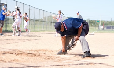 An umpire is cleaning home plate on a baseball or softball field. Behind him, a group of players in white and red uniforms are gathered, with some interacting with a coach. The field is enclosed by a chain-link fence, and there is a grassy area and trees in the background.