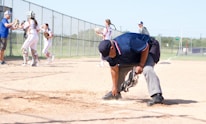 An umpire is cleaning home plate on a baseball or softball field. Behind him, a group of players in white and red uniforms are gathered, with some interacting with a coach. The field is enclosed by a chain-link fence, and there is a grassy area and trees in the background.