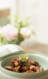 A bowl of cooked food, featuring pieces of meat and vegetables, garnished with fresh green herbs. The dish is presented in a pastel-colored bowl, with a soft and blurred background of flowers, providing a warm and inviting atmosphere.