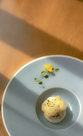 A beautifully plated dish featuring a delicate piece of sushi or nigiri with creamy yellow topping, garnished with small black seeds and surrounded by a sprig of microgreens and an edible flower. The dish is presented on a minimalist, light gray plate, casting soft shadows across the wooden table.