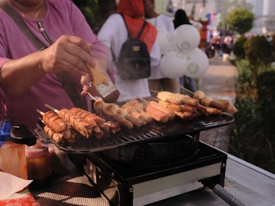 A person with a purple shirt is grilling skewered sausages and meats on a hot barbecue. The grill is placed on a metal table, and the person is using a brush to apply sauce onto the skewers. Various condiments and bottles are nearby. In the background, several people, including one wearing a red hijab, are visible, along with some white balloons.