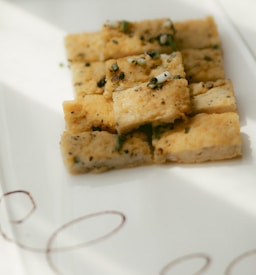 Squares of herb-infused bread or focaccia are neatly arranged on a white plate. The bread appears to have a golden-brown crust with visible pieces of herbs and possibly green onions sprinkled on top.