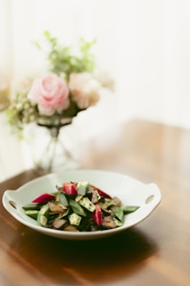 A stylishly arranged dish featuring sliced vegetables, including okra and red peppers, served in a white bowl. The background showcases a blurred vase of soft pink and white flowers on a wooden table, adding a touch of elegance and warmth.