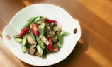 A white plate contains a colorful dish featuring sliced green okra, red chili peppers, and cooked mushrooms. The vegetables are artfully arranged and the composition appears fresh and appetizing. The plate is set on a wooden surface with natural light creating soft shadows.