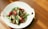 A white plate contains a colorful dish featuring sliced green okra, red chili peppers, and cooked mushrooms. The vegetables are artfully arranged and the composition appears fresh and appetizing. The plate is set on a wooden surface with natural light creating soft shadows.