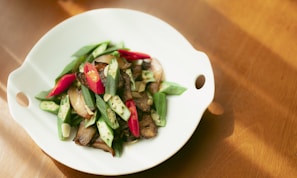 A white plate contains a colorful dish featuring sliced green okra, red chili peppers, and cooked mushrooms. The vegetables are artfully arranged and the composition appears fresh and appetizing. The plate is set on a wooden surface with natural light creating soft shadows.