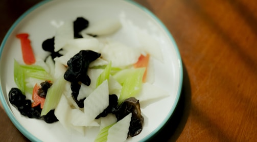 A plate with a mix of sliced vegetables, including white radish, black fungus, and green and red peppers. The dish is served on a white plate with a blue rim, placed on a wooden surface.