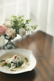 A white bowl sits on a wooden table, filled with a creamy soup garnished with what appears to be chicken pieces, herbs, and possibly mushrooms. In the background, there is a delicate bouquet of flowers in soft colors like pink, white, and green, placed in a glass vase. Natural light filters through sheer curtains, casting gentle shadows.