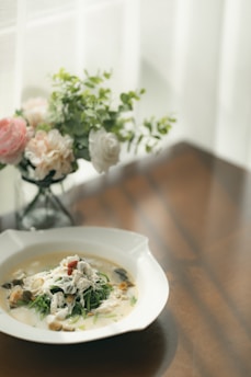 A white bowl sits on a wooden table, filled with a creamy soup garnished with what appears to be chicken pieces, herbs, and possibly mushrooms. In the background, there is a delicate bouquet of flowers in soft colors like pink, white, and green, placed in a glass vase. Natural light filters through sheer curtains, casting gentle shadows.