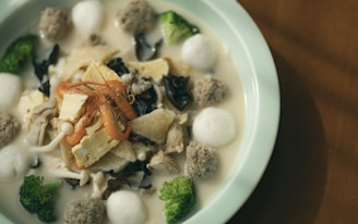 A dish with various ingredients including shrimp, tofu, black fungus, broccoli, meatballs, and soup in a light, creamy broth. Presented in a round ceramic bowl on a wooden table.
