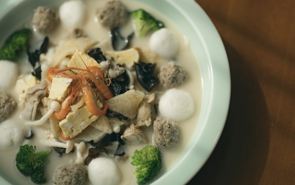 A dish with various ingredients including shrimp, tofu, black fungus, broccoli, meatballs, and soup in a light, creamy broth. Presented in a round ceramic bowl on a wooden table.