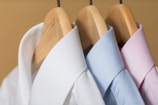 Three neatly pressed collared shirts hang on wooden hangers. The shirts are arranged closely together with a white shirt on the left, a light blue shirt in the center, and a light pink shirt on the right. The background is a soft beige color, creating a neutral and soothing setting.