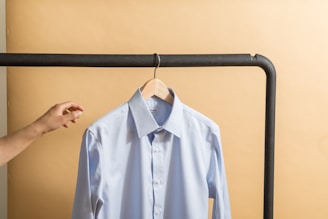 A neatly pressed light blue shirt is hanging on a wooden hanger, supported by a black metal clothing rack. A hand is reaching towards the shirt against a beige background.