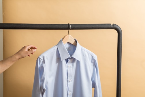 A stylish men's navy blue casual shirt displayed on a wooden hanger against a soft pink background.