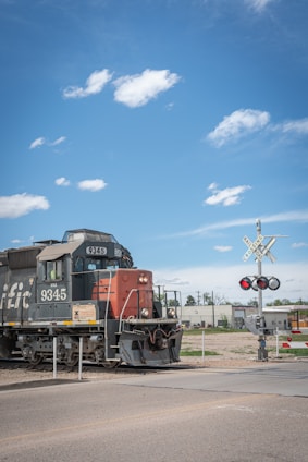 A freight train with the number 9345 is stationed on a railway track next to a railroad crossing sign. The locomotive has a mix of black and red colors. There are industrial buildings in the background along with some sparse vegetation. The sky is clear with a few scattered clouds.