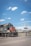 A freight train with the number 9345 is stationed on a railway track next to a railroad crossing sign. The locomotive has a mix of black and red colors. There are industrial buildings in the background along with some sparse vegetation. The sky is clear with a few scattered clouds.