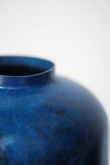 A close-up view of a deep blue ceramic vase with a slightly textured surface. The upper edge of the vase is visible, and the background is a soft white, enhancing the simplicity and elegance of the design.