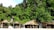 A tropical beach scene with thatched roof huts set against a backdrop of dense, lush greenery and rugged rock formations. The foreground is sandy, while the greenery includes a variety of trees and shrubs.