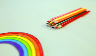 A set of colored pencils is neatly arranged on a light background next to a hand-drawn rainbow. The rainbow is vibrant, displaying a full spectrum of colors from red to purple.