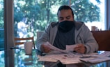 A man wearing a dark face mask and casual clothing sits at a table covered in papers. He is focused on examining a document while a white mug rests nearby. The background reveals large windows displaying a lush green outdoor scene, suggesting a peaceful setting.