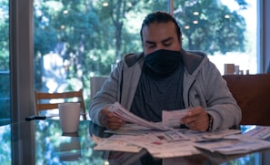 A man wearing a dark face mask and casual clothing sits at a table covered in papers. He is focused on examining a document while a white mug rests nearby. The background reveals large windows displaying a lush green outdoor scene, suggesting a peaceful setting.