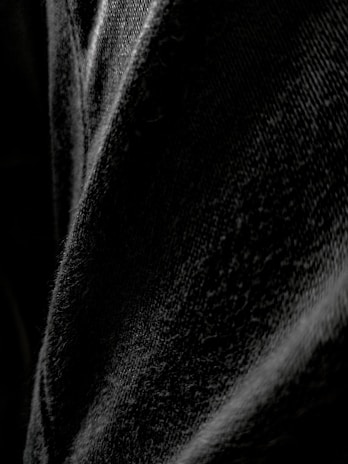 A close-up view of black denim fabric, highlighting the texture and weave of the material with varying shades of black and gray.