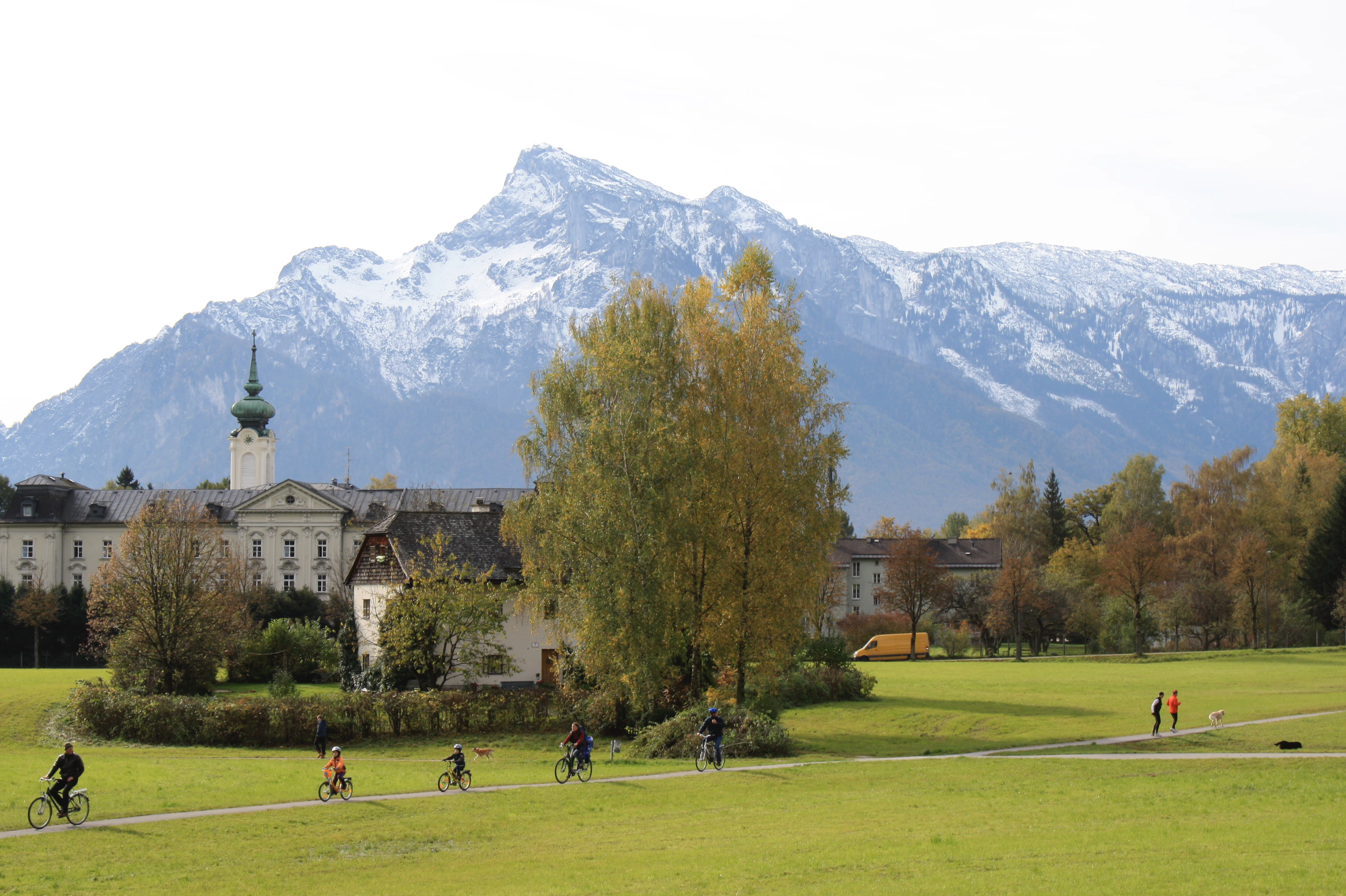 Cyclists ride through green parkland below a towering Alpine peak on Salzburg's outskirts, autumn trees in the foreground.