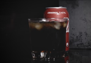 A cold beverage can with the text 'original taste' is partially visible, placed behind a glass filled with a dark soda and ice cubes. The background is dark, highlighting the condensation on the can and the drink's refreshing appearance.