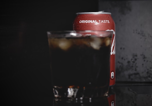 A cold beverage can with the text 'original taste' is partially visible, placed behind a glass filled with a dark soda and ice cubes. The background is dark, highlighting the condensation on the can and the drink's refreshing appearance.