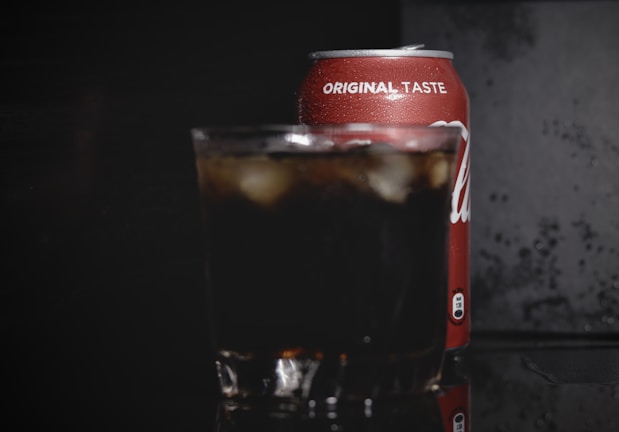 A cold beverage can with the text 'original taste' is partially visible, placed behind a glass filled with a dark soda and ice cubes. The background is dark, highlighting the condensation on the can and the drink's refreshing appearance.