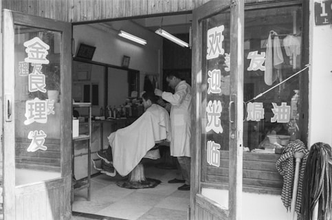 A vintage barbershop scene with a barber cutting a customer's hair. The shop interior features a simple setup with mirrors and shelves lined with various grooming products. The entrance door has Chinese characters on it. The atmosphere is classic, reflecting an old-fashioned style with wooden elements and a tiled floor.