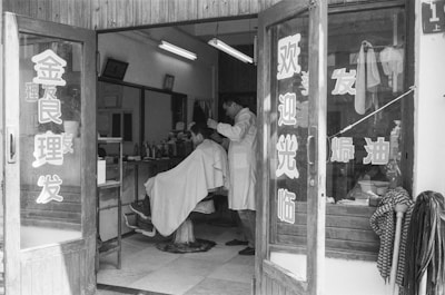 A vintage barbershop scene with a barber cutting a customer's hair. The shop interior features a simple setup with mirrors and shelves lined with various grooming products. The entrance door has Chinese characters on it. The atmosphere is classic, reflecting an old-fashioned style with wooden elements and a tiled floor.