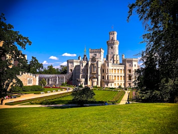 A grand, medieval-style castle sits prominently in a lush, green landscape under a clear blue sky. The castle is adorned with towers and intricate architectural details, surrounded by manicured lawns and gardens. Visitors are visible walking along the pathways and relaxing on benches, contributing to a tranquil and picturesque scene.