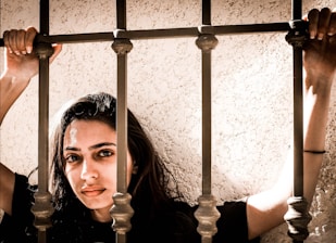 A woman with long brown hair is holding onto metal bars with both hands. Her expression is serious and she is positioned against a textured beige wall. The lighting creates a dramatic shadow effect.