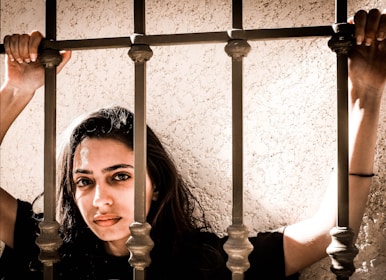 A woman with long brown hair is holding onto metal bars with both hands. Her expression is serious and she is positioned against a textured beige wall. The lighting creates a dramatic shadow effect.