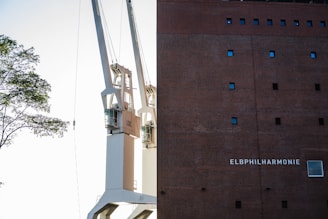 Two large cranes are positioned next to a tall brick building with small square windows. The building has the name 'Elbphilharmonie' displayed on it. A tree branch with green leaves is visible on the left side.