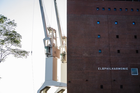 Two large cranes are positioned next to a tall brick building with small square windows. The building has the name 'Elbphilharmonie' displayed on it. A tree branch with green leaves is visible on the left side.