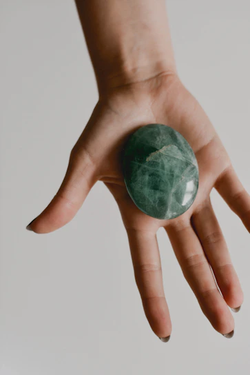 Close-up of a hand holding a polished holy stone with subtle gold veins, against a soft off-white background.