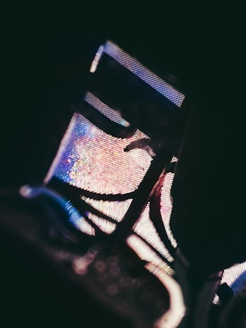 A close-up image focusing on a section of a light structure, possibly a digital screen or sign. The image displays pixels in vibrant colors, including blue, pink, and purple, creating abstract patterns. The view is slightly obscured by shadows and metallic elements, giving it an artistic, high-contrast appearance.