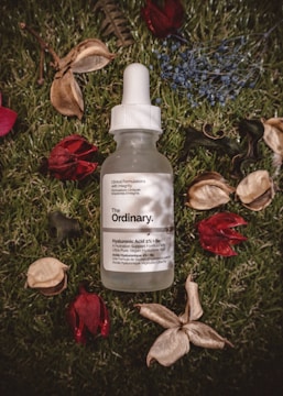 A bottle with a dropper labeled 'The Ordinary' containing Hyaluronic Acid 2% + B5 is positioned on a grassy surface. Around the bottle, there are scattered dried flowers and leaves, adding a natural and rustic touch to the scene.